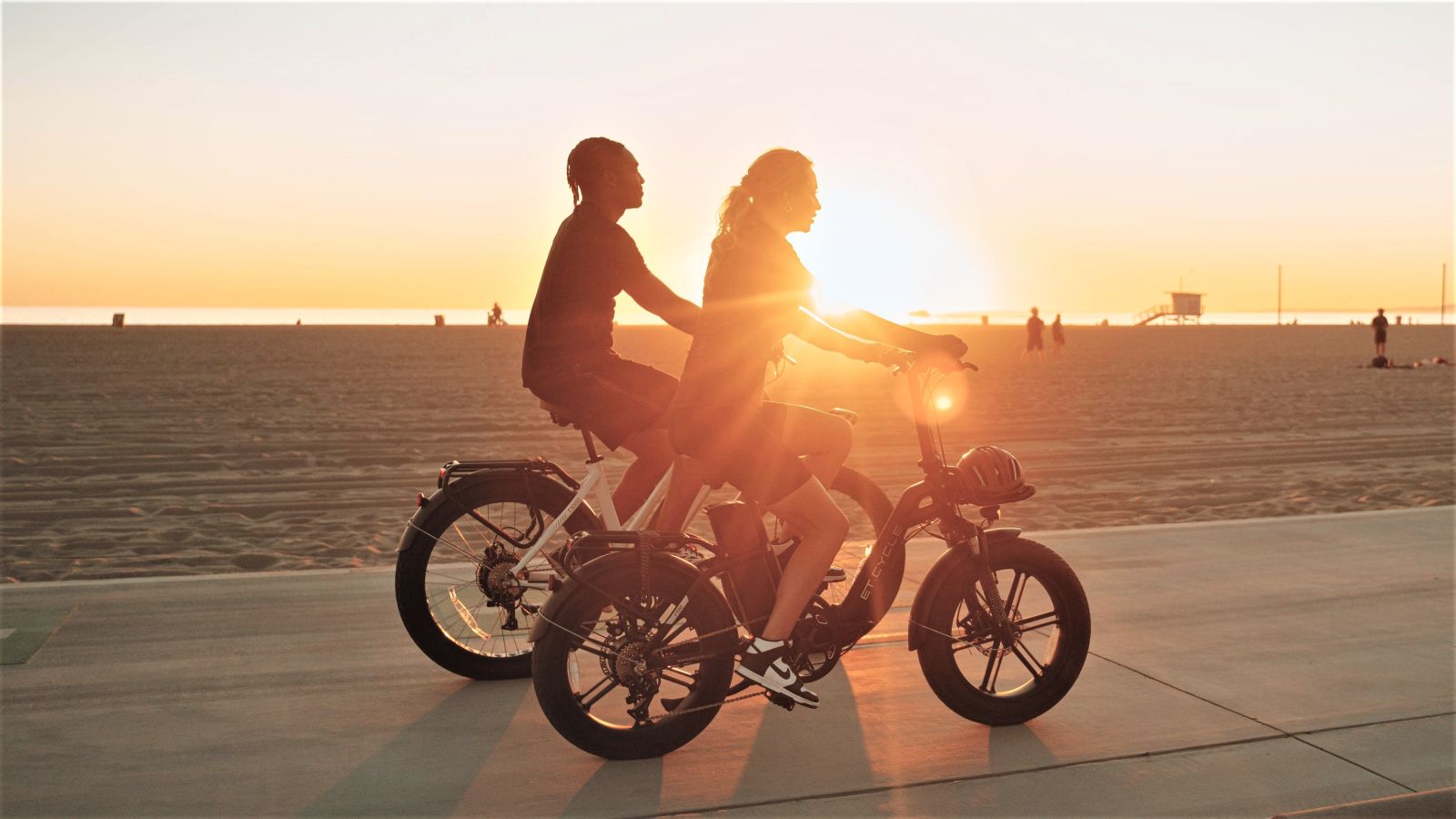 Two riders on NCM e-bikes on a beach path at sunset